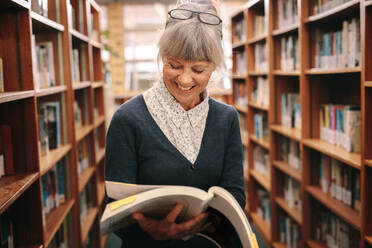 Cheerful senior woman standing in a library looking at a text book. Senior woman checking for reference books in a university library. - JLPSF22276