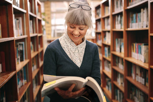 Fröhliche ältere Frau, die in einer Bibliothek steht und sich ein Lehrbuch ansieht. Ältere Frau, die in einer Universitätsbibliothek nach Nachschlagewerken sucht. - JLPSF22276