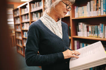 Side view of an elderly woman standing in a library flipping pages of a text book. Senior woman checking reference books standing in a university library. - JLPSF22278
