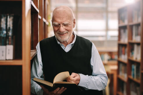 Fröhlicher älterer Mann, der in einer Bibliothek steht und sich ein Lehrbuch ansieht. Älterer Mann, der in einer Universitätsbibliothek nach Nachschlagewerken sucht. - JLPSF22279