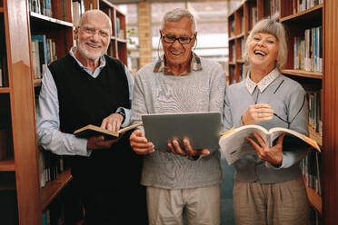 Lächelnde ältere Männer und Frauen stehen in einer Bibliothek mit Büchern und Laptop. Glückliche Senioren genießen das Lesen und Lernen in der Bibliothek. - JLPSF22280