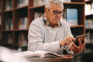 Älterer Lernender, der ein Buch in einer Bibliothek liest und dabei auf einen Tablet-PC schaut. Älterer Mann, der in einer Bibliothek sitzt und ein Buch liest. - JLPSF22283