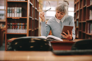 Senior woman using a tablet pc for reference while reading a book in library. Woman sitting in a library reading a book. - JLPSF22284