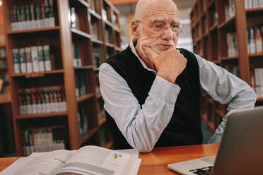 Senior man sitting in a library reading on a laptop computer with hand to his chin. Elderly man sitting in classroom and learning with a laptop and books on the table. - JLPSF22286