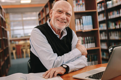 Lächelnder älterer Mann sitzt in einer Bibliothek mit seinen Büchern und Laptop auf dem Tisch. Fröhlicher älterer Mann sitzt in der Bibliothek und lernt. - JLPSF22287