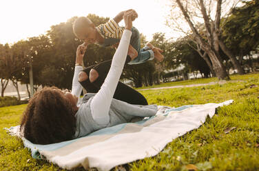 Mutter spielt mit ihrem Sohn im Park. Afrikanische Frau liegt im Park auf einer Decke und hebt den Jungen auf ihre Füße. - JLPSF22297