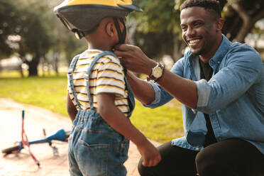Ein lächelnder Mann hilft seinem Sohn beim Tragen eines Fahrradhelms im Park. Ein Junge macht sich mit einem Fahrradhelm bereit, mit dem Radfahren zu beginnen. - JLPSF22302
