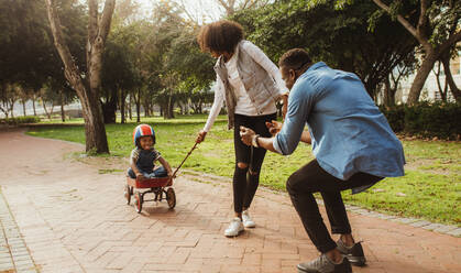 Boy sitting in a small wagon pulled by his mother, while father standing with his arms outstretched. Young family on enjoying at park. - JLPSF22308