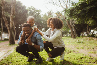 Mann, der seinen Sohn auf dem Rücken trägt, mit Frau, die ihn im Park unterstützt. Glückliche junge afrikanische Familie, die sich im Park vergnügt. - JLPSF22310