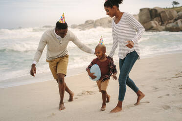 Young african family wearing party hat running along the beach. Couple with son running by the sea shore. - JLPSF22319