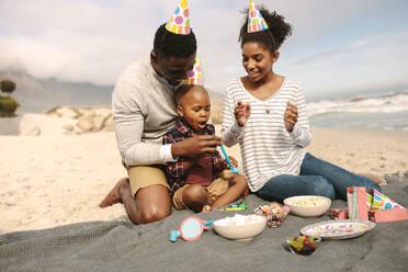Junger Vater und Mutter feiern den Geburtstag ihres Sohnes. Die Familie trägt Partyhüte und feiert den Geburtstag des Sohnes am Strand. - JLPSF22323