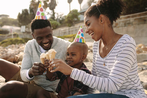 Afrikanische Familie mit Partyhüten isst Eis am Strand. Mann und Frau mit Sohn genießen Eis essen. Familie genießt den Geburtstag des Sohnes mit Eis im Freien. - JLPSF22326