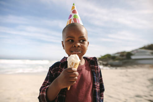 Afrikanischer Junge mit Partyhut isst Eistüte am Strand. Geburtstagskind isst Eis am Meer. - JLPSF22327