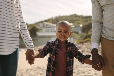 Junge hält seine Mutter und seinen Vater an den Händen, während er am Strand spazieren geht. Junge mit seinen Eltern am Meer. - JLPSF22330