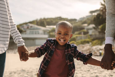 Lächelnder Junge, der Vater und Mutter am Strand an den Händen hält. Familie geht am Strand entlang. - JLPSF22331
