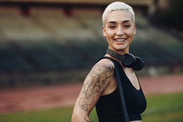 Fröhliche Fitness-Frau in einem Leichtathletik-Stadion mit ihrer Tasche und drahtlosen Kopfhörern. Seitenansicht einer Athletin in einem Leichtathletik-Stadion. - JLPSF22344