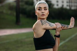 Woman athlete with tattoo on her arm doing stretching exercises in a ground. Portrait of a woman in fitness clothes doing warm up exercises. - JLPSF22349