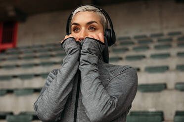 Frau im Sweatshirt steht in einem Stadion und hört Musik. Fitness-Frau entspannt sich nach dem Training und hört Musik über Kopfhörer. - JLPSF22364