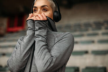 Fitness-Frau entspannt sich nach dem Training und hört Musik über Kopfhörer in einem Stadion. Porträt einer Frau in Jacke auf der Tribüne eines Stadions. - JLPSF22365