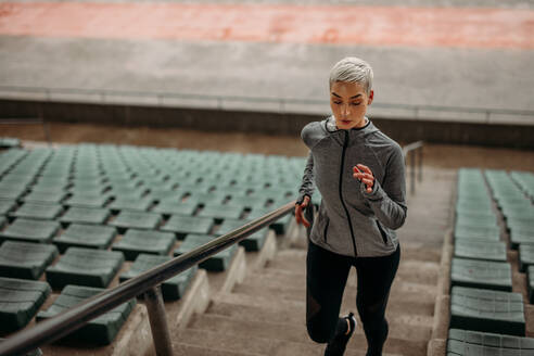 Female athlete running up the stairs of a stadium. Woman in fitness wear doing workout running on stairs beside the seats of a stadium. - JLPSF22367