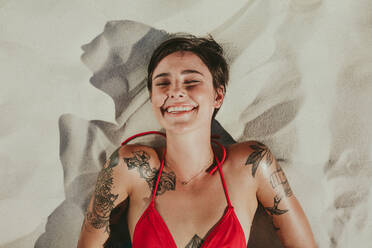 Close up top view of a smiling woman in bikini lying on beach sand. Close up of a woman with tattoos over her body relaxing with closed eyes on beach. - JLPSF22400