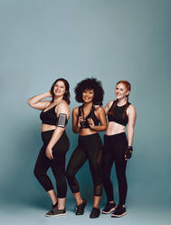 Gruppe von verschiedenen Frauen in Sportkleidung, die vor einem grauen Hintergrund zusammenstehen. Drei Frauen unterschiedlichen Gewichts in Fitnesskleidung. - JLPSF22407