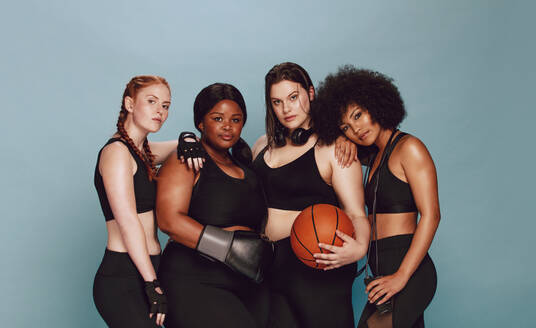 Multiethnische Frauen in Sportkleidung stehen mit Basketball, Boxhandschuhen und Springseil zusammen. Unterschiedliche Gruppe von Frauen mit Sportgeräten schaut in die Kamera vor grauem Hintergrund. - JLPSF22422