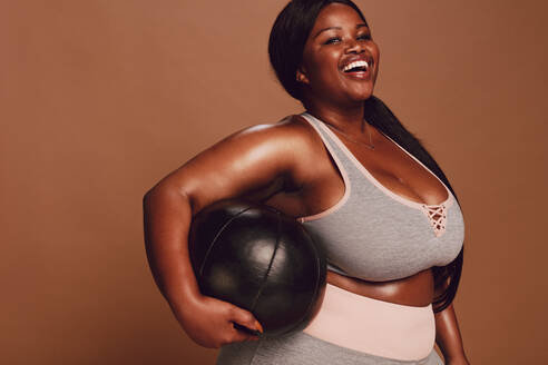 Fröhliche große afrikanische Frau mit einem Medizinball auf braunem Hintergrund. Plus Size Frau in Sportkleidung schaut in die Kamera und lacht. - JLPSF22444