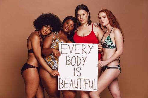 Diverse Gruppe von Frauen mit einem Schild von jedem Körper ist schön gegen braunen Hintergrund. Verschiedene Größe Frauen in Bikinis mit einem Plakat. - JLPSF22458