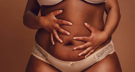 Cropped shot of black female in underwear holding her fat belly. Plus size woman in lingerie holding her belly with both hands on brown background. - JLPSF22472