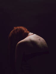 Rückansicht einer sehr dünnen rothaarigen Frau, die sich für ihren Körper schämt. Eine untergewichtige Frau fühlt sich vor einem dunklen Hintergrund traurig. - JLPSF22478