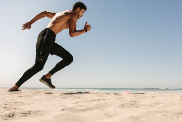 Mann beim Fitnesstraining am Strand an einem sonnigen Tag. Seitenansicht eines sportlichen Mannes, der zu einem Sprint am Strand ansetzt. - JLPSF22495