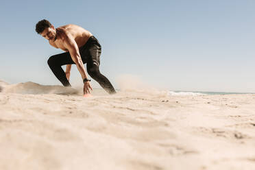 Athletic man bending low to touch the mark while running on beach. Man doing fitness work out at a beach on a sunny day. - JLPSF22496
