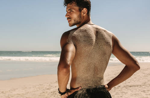 Rückansicht eines Mannes mit nacktem Oberkörper, der während des Trainings am Strand steht und Sand auf seinem Rücken hat. Sportlicher Mann, der am Strand steht und die Hände auf die Hüfte legt, während er ein Fitnesstraining absolviert. - JLPSF22504