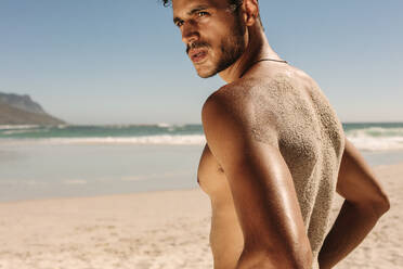 Seitenansicht eines Mannes mit nacktem Oberkörper, der während des Trainings am Strand steht und Sand auf seinem Rücken hat. Athletischer Mann, der am Strand steht und sich beim Fitnesstraining entspannt. - JLPSF22505