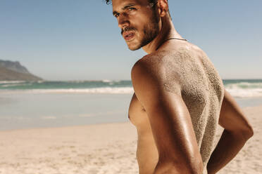 Side view of a bare chested man standing on a beach during workout with sand attached to his back. Athletic man standing on beach relaxing while doing fitness workout. - JLPSF22505