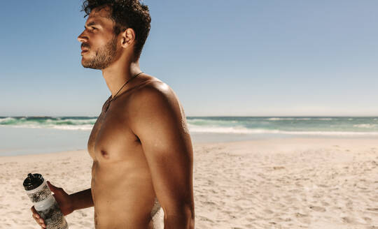 Side view of a bare chested man standing on a beach during workout holding a water bottle. Athletic man standing on beach taking a break while doing fitness workout. - JLPSF22506