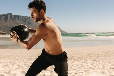 Ein Mann macht ein Fitnesstraining am Strand mit einem Medizinball. Ein athletischer Mann mit nacktem Oberkörper macht Drehungen mit einem Medizinball. - JLPSF22511