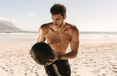 Ein Mann macht ein Fitnesstraining am Strand mit einem Medizinball. Ein athletischer Mann mit nacktem Oberkörper macht Drehungen mit einem Medizinball. - JLPSF22514