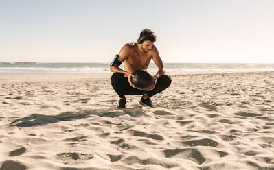 Ein Mann macht ein Fitnesstraining am Strand mit einem Medizinball. Ein athletischer Mann mit nacktem Oberkörper macht Kniebeugen und hält einen Medizinball. Er trägt drahtlose Kopfhörer und ein Mobiltelefon an der Armbinde. - JLPSF22516