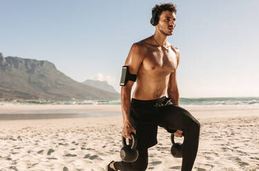 Man doing fitness workout at a beach using kettlebells. Bare chested athletic man doing exercise with kettlebells wearing wireless headphones and mobile phone fixed to armband. - JLPSF22517