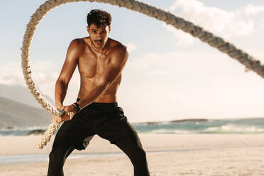 Man doing fitness workout at a beach on a sunny day. Bare chested man doing workout using a battle rope on the beach. - JLPSF22525