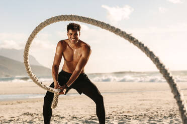 Mann beim Fitnesstraining am Strand an einem sonnigen Tag. Mann mit nacktem Oberkörper beim Workout mit einem Battle Rope am Strand. - JLPSF22526