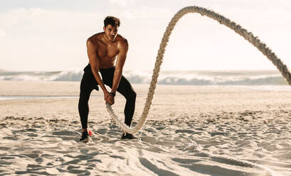 Mann beim Fitnesstraining am Strand an einem sonnigen Tag. Mann mit nacktem Oberkörper beim Workout mit einem Battle Rope am Strand. - JLPSF22527