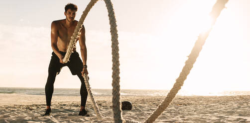 Mann beim Fitnesstraining am Strand an einem sonnigen Tag. Mann mit nacktem Oberkörper beim Workout mit zwei Battle Ropes am Strand. - JLPSF22533