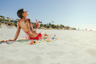 Frau im Badeanzug, die auf einem Handtuch sitzt und sich am Strand sonnt und eine Flasche in der Hand hält. Frau mit Tattoos auf ihrem Körper im Urlaub, die sich an einem sonnigen Tag am Strand entspannt. - JLPSF22541