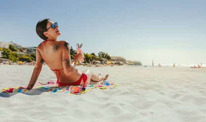 Lächelnde Frau im Bikini, die auf einem Handtuch sitzt und sich am Strand sonnt und eine Flasche in der Hand hält. Frau mit Sonnenbrille und Tattoos auf ihrem Körper im Urlaub, die sich an einem sonnigen Tag am Strand entspannt. - JLPSF22542