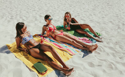 Women relaxing on beach with drinks in hand on a sunny day. Three girlfriends lying on beach in bikini sunbathing while enjoying their drinks. - JLPSF22548