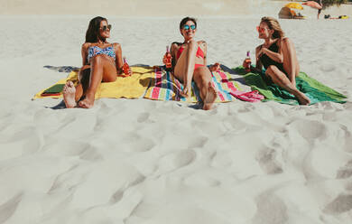 Three girlfriends sitting on beach in bikini sunbathing while enjoying their drinks. Smiling women wearing sunglasses relaxing on beach with drinks in hand on a sunny day. - JLPSF22549