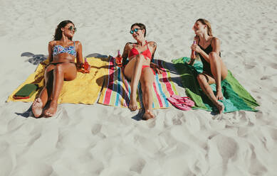 Three girlfriends sitting on beach in bikini sunbathing while enjoying their drinks. Smiling women wearing sunglasses relaxing on beach with drinks in hand on a sunny day. - JLPSF22550